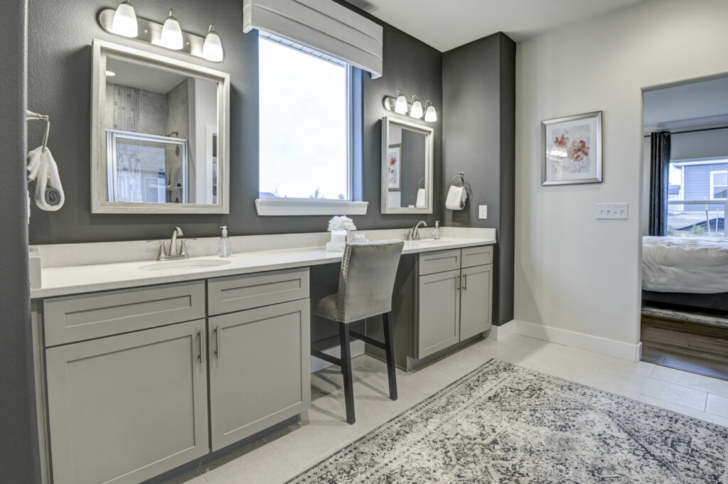 Double-sink bathroom vanity with gray cabinets, two mirrors, window, and a chair at the center vanity area.
