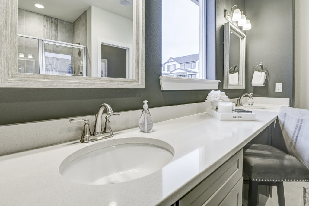 Close-up of a bathroom countertop with under-mount sink, faucet, soap dispenser, and window reflection.