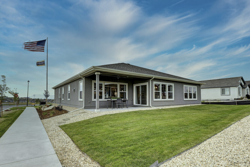 Single-story gray house with covered patio, lawn, and flagpole under a cloudy blue sky.