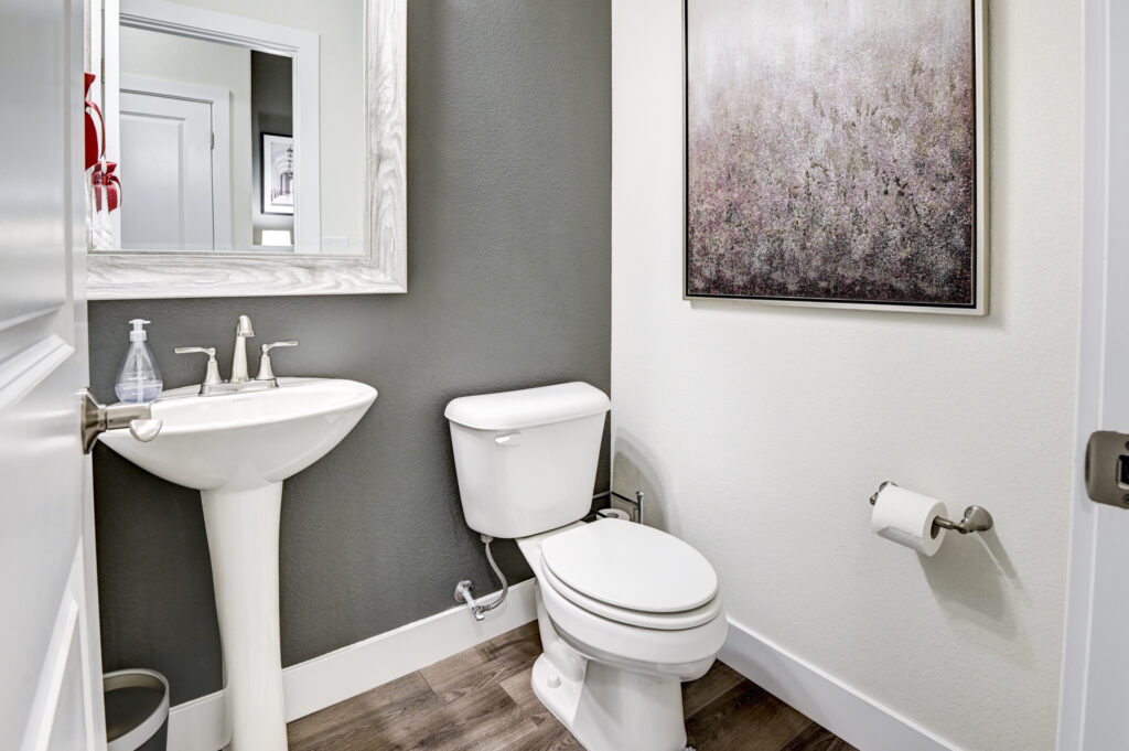 Small half-bath with pedestal sink, toilet, framed mirror and wall art on white and gray walls.