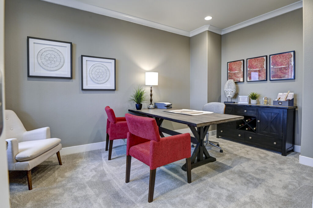Carpeted office with a wooden desk, two red upholstered chairs, and artwork on neutral walls.