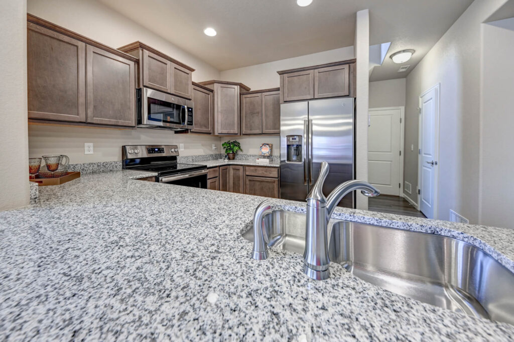 Close-up of granite countertop and sink with faucet, kitchen cabinets and stainless fridge beyond.