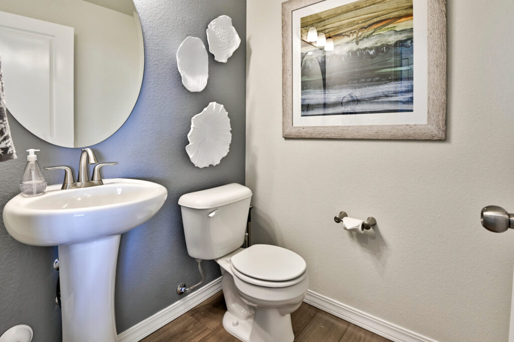 Small residential powder room with pedestal sink, toilet, round mirror, and framed artwork.