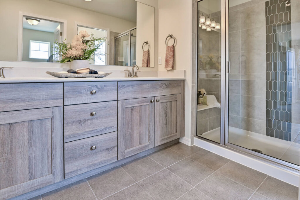 Close view of gray wood vanity cabinets, drawers, tiled floor and shower door.