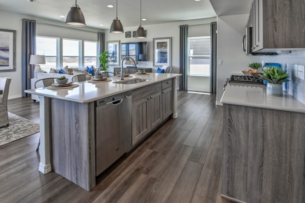 Open kitchen with large island, stainless dishwasher, pendant lights, and adjacent living area.
