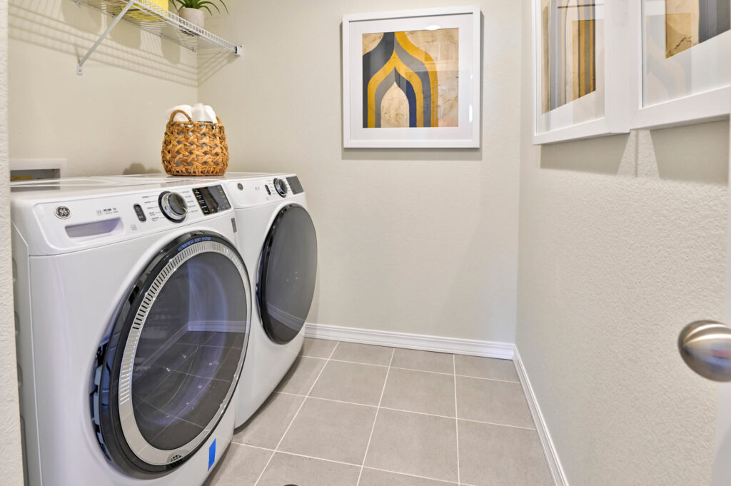 Laundry room with front-loading washer and dryer, tiled floor, wall shelving, and framed artwork.