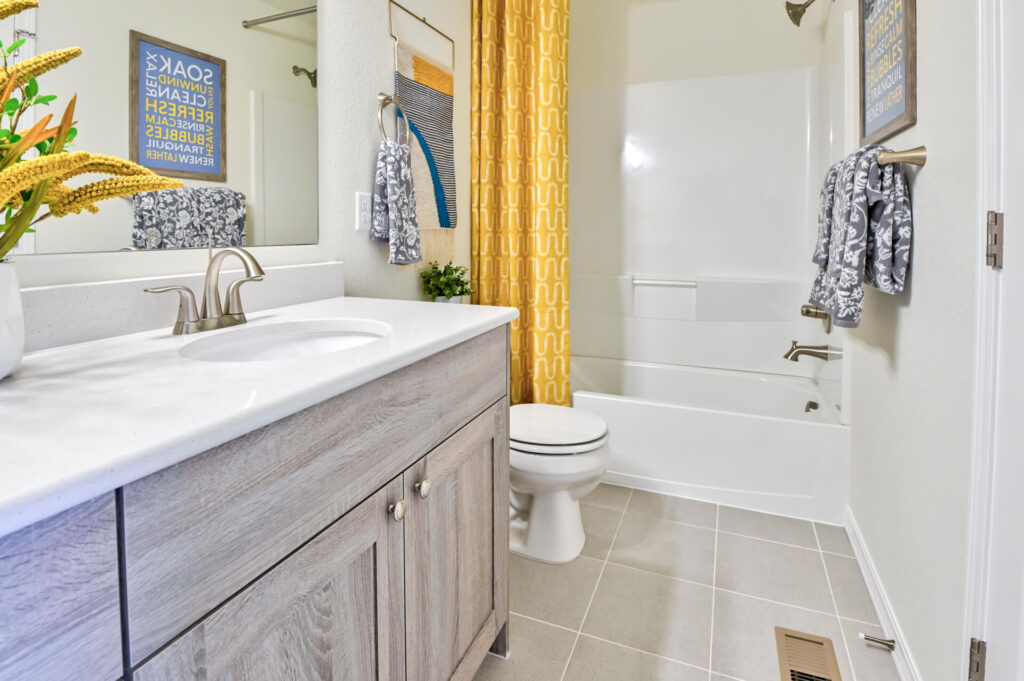 Bathroom with single sink vanity, shower-tub, yellow patterned shower curtain and neutral tile floor.