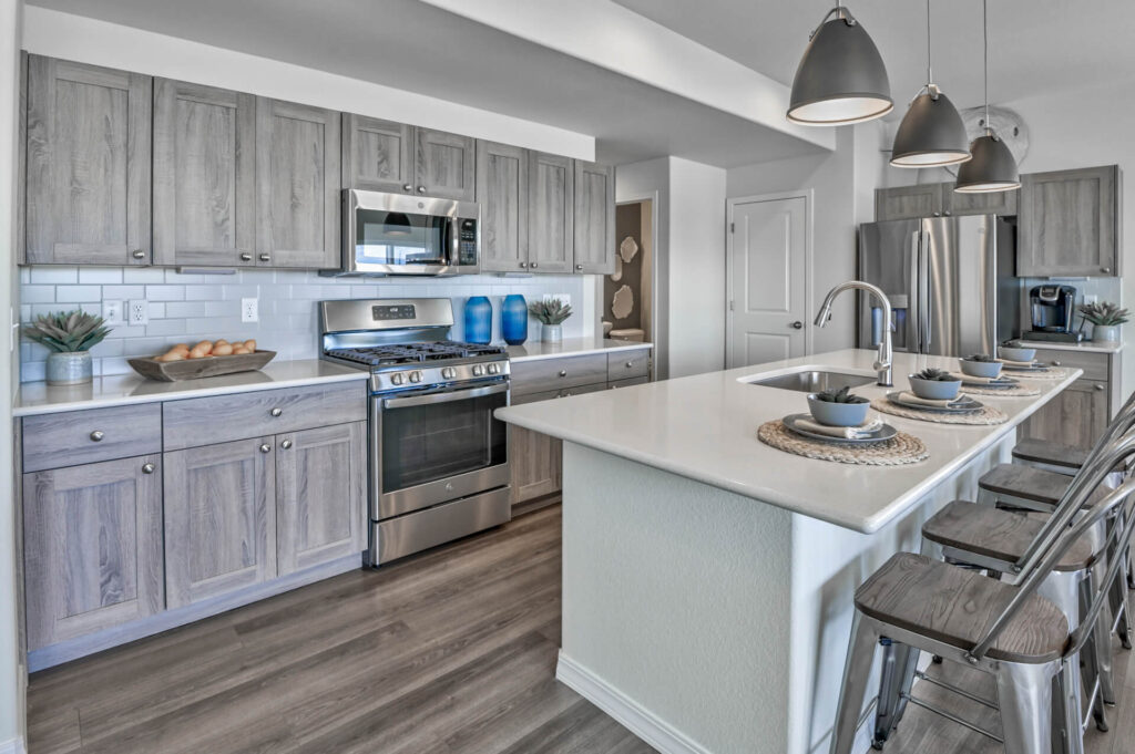 Bright modern kitchen with long island, stainless appliances, gray wood cabinets and pendant lights.