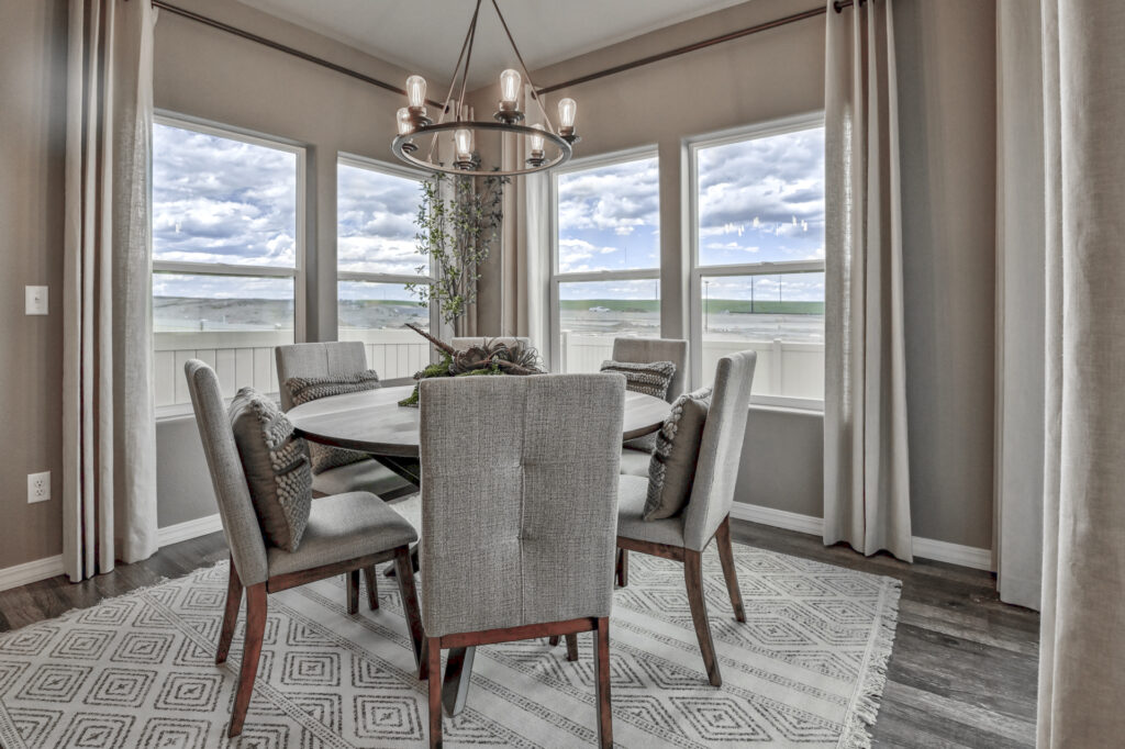 Dining area with round table, six upholstered chairs and bay of three windows under a chandelier.