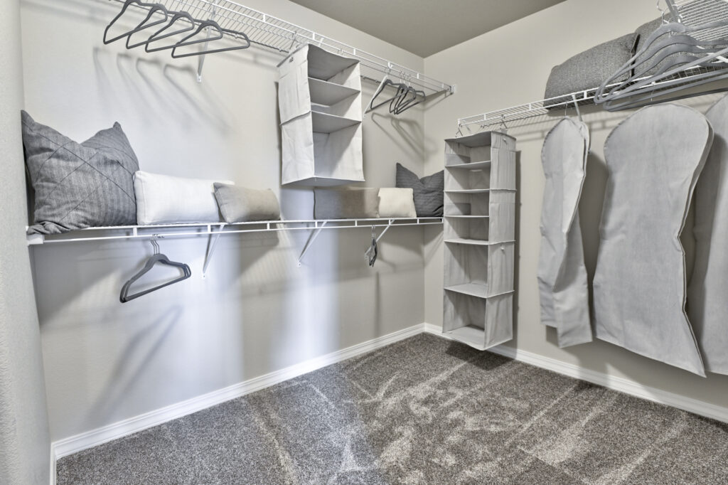 Empty walk-in closet with carpet, white wire shelving, hanging organizers and gray pillows.