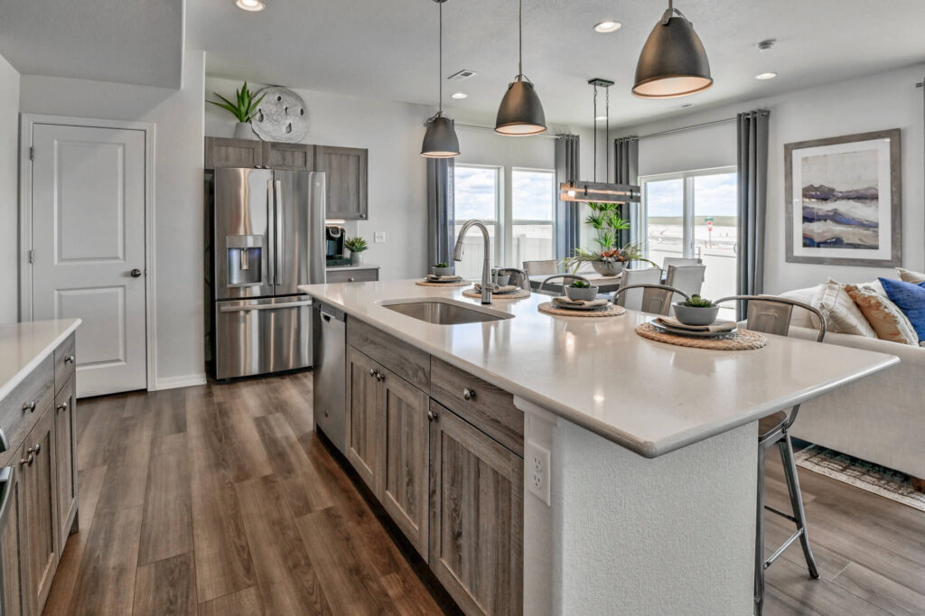 Kitchen island view toward dining area with stainless fridge, sink in island and three pendant lamps.