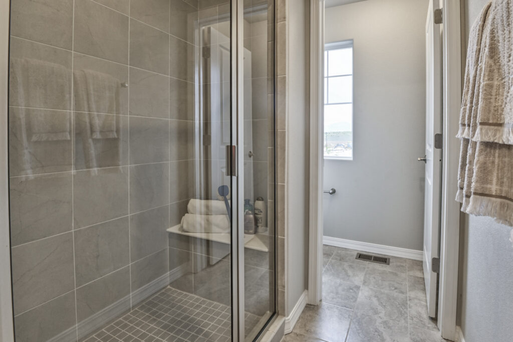 Tiled shower with glass doors, built-in bench holding towels and toiletries, and adjacent toilet alcove.