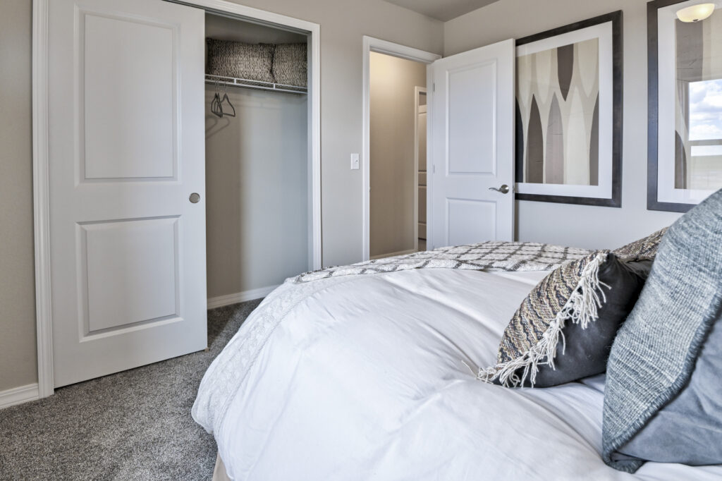 Bedroom corner showing bed linens, an open closet door, entry door, and framed wall art.