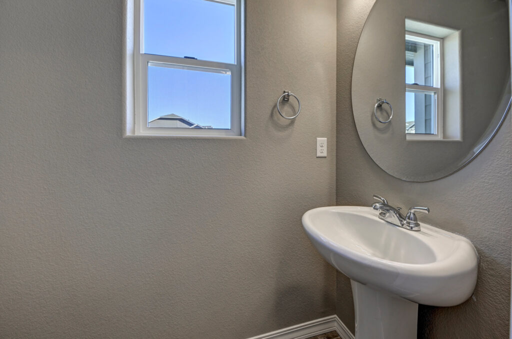 Small powder room with pedestal sink, round mirror, and a window letting in daylight.
