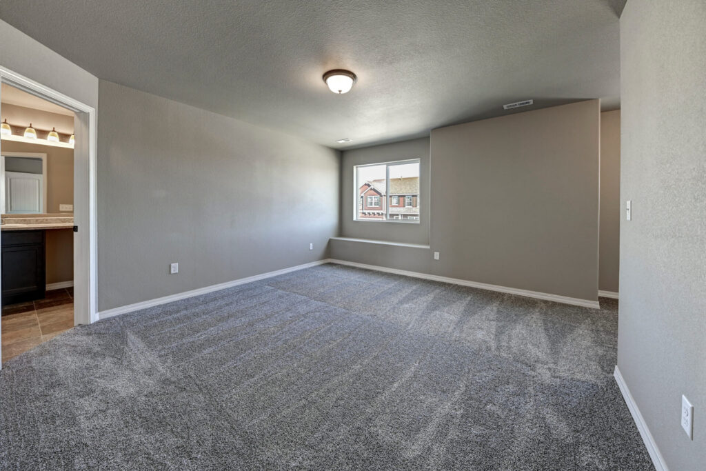 Empty carpeted bedroom with a window, neutral walls, and an adjacent bathroom doorway.