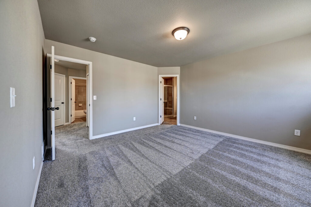 Medium-sized carpeted bedroom with open doors to bathroom and closet, neutral walls and ceiling light.