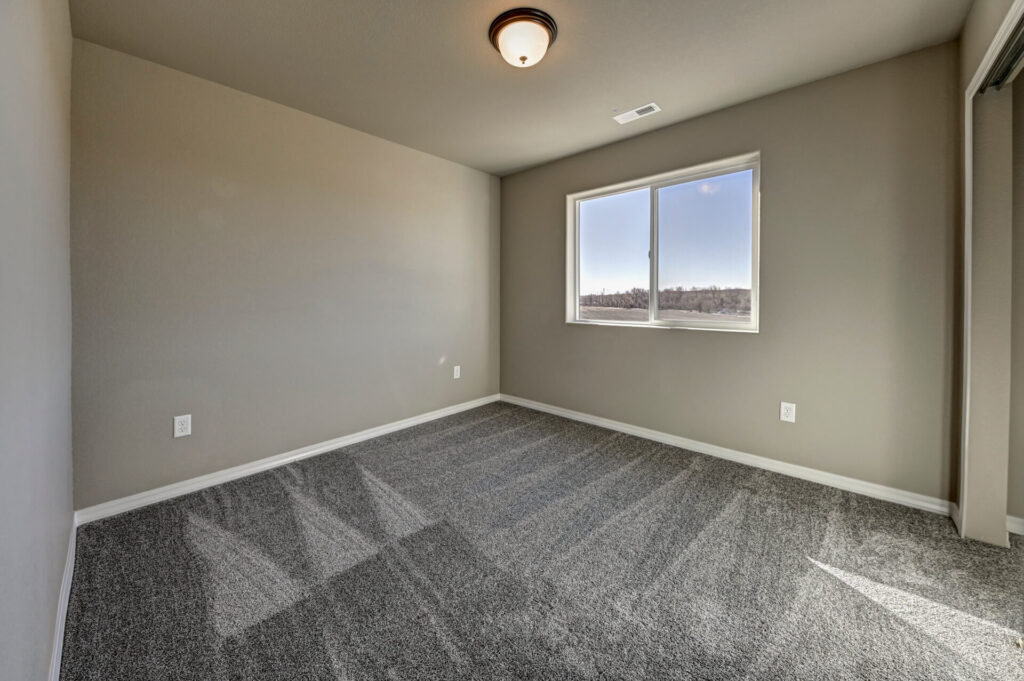Empty bedroom with window, neutral walls, and freshly vacuumed carpet with visible pattern.