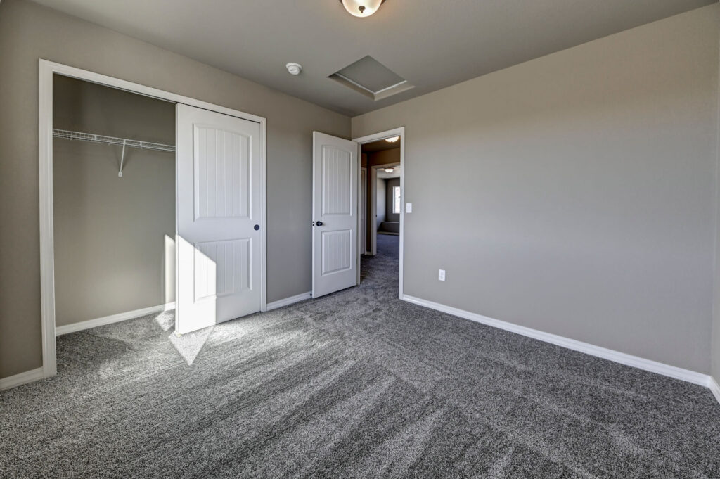 Empty carpeted bedroom with open closet and white paneled doors, neutral walls and ceiling light.