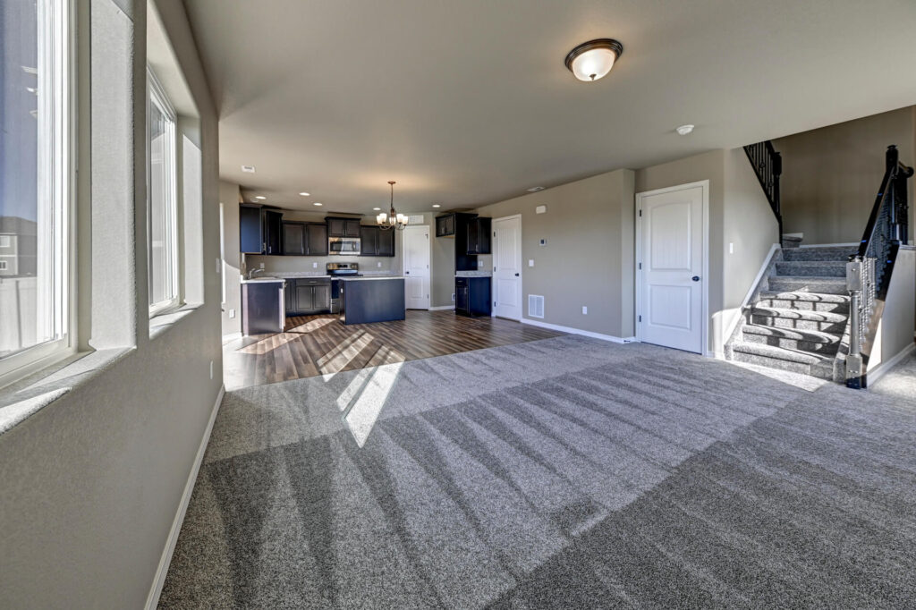 Living room view toward kitchen with carpeted floor, sunlight patterns, and staircase on the right.