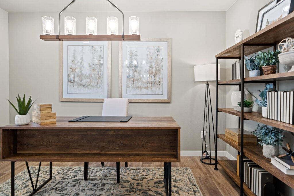 Home office setup with wooden desk, patterned rug, shelving and framed artwork on wall.