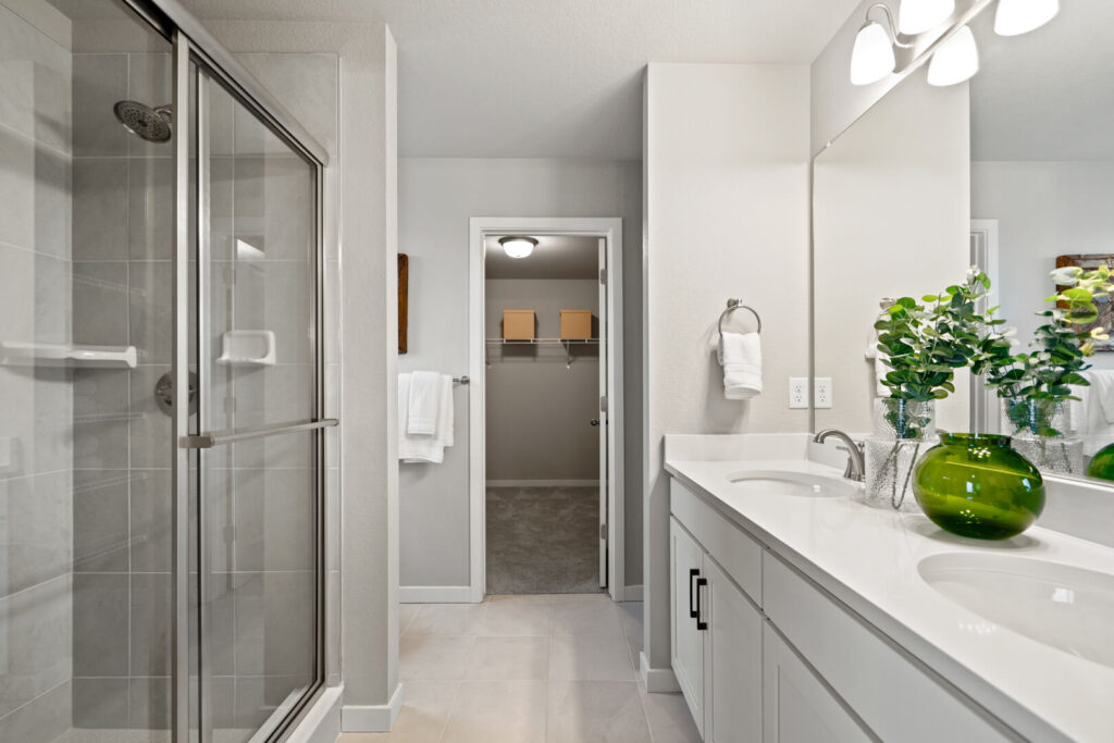 Bathroom vanity with double sinks, large mirror, and a glass-enclosed shower next to an open doorway.