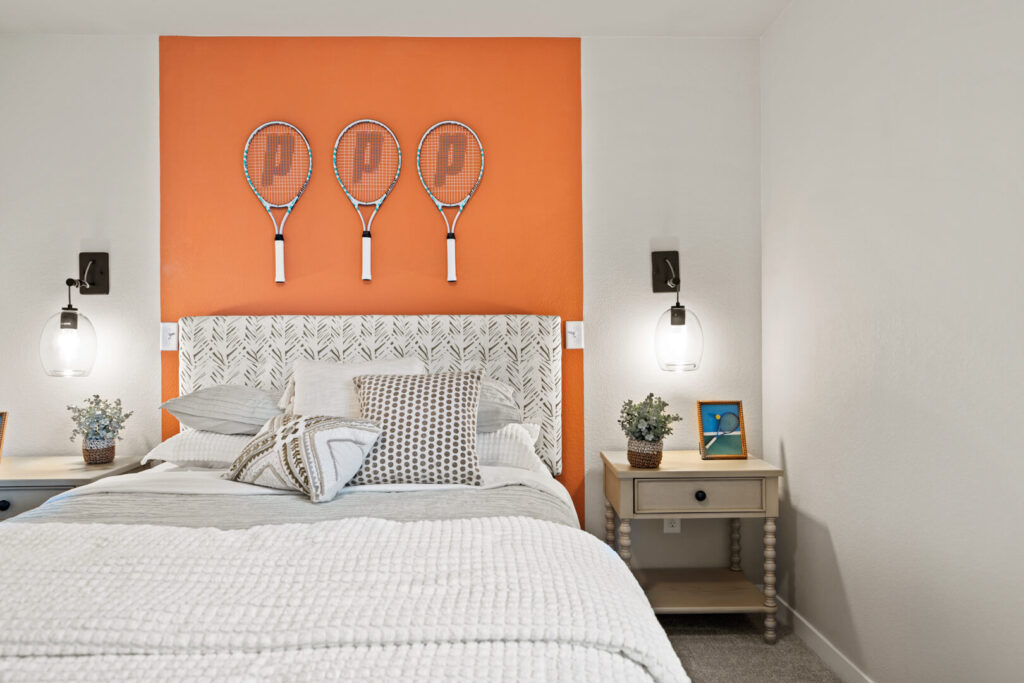 Bed with patterned headboard, orange accent wall and three tennis racquets mounted above.