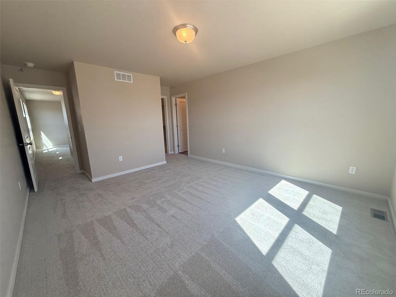 bedroom with neutral walls, wall vent above short partition and sunlight window patch on carpet